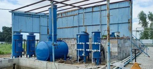 Top Rated Residential STP Plant Manufacturers for Residential Complexes Industrial water treatment setup with multiple blue cylindrical filtration tanks, connected piping, and a horizontal pressure vessel inside a partially open metal shed.
