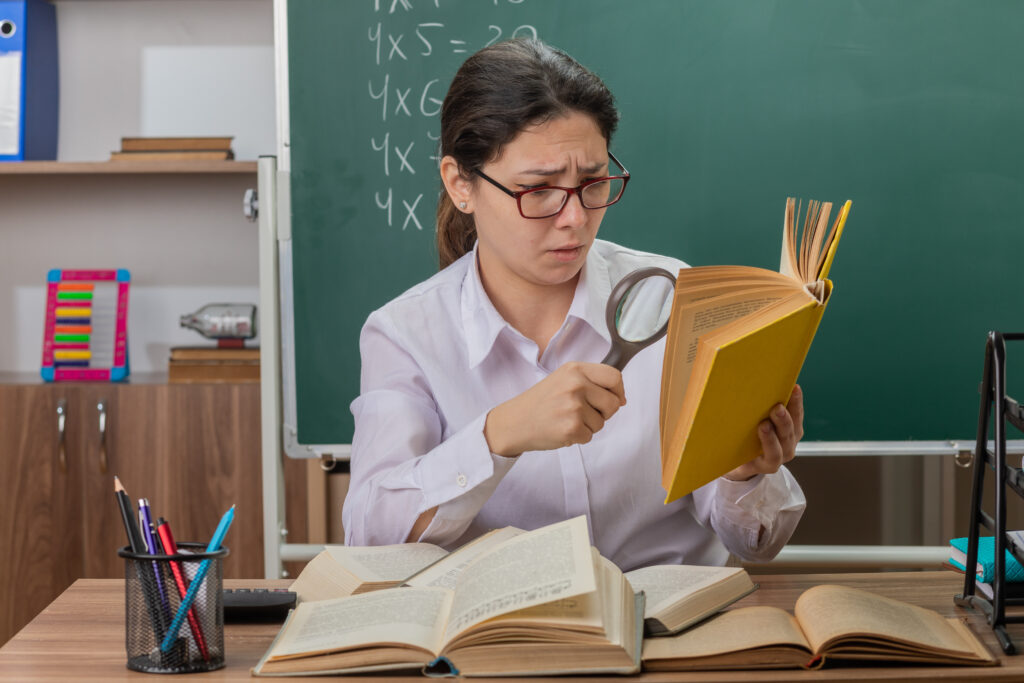 young woman teacher wearing glasses looking at book through magnifying glass being confused and displeased sitting at school desk in front of blackboard in classroom